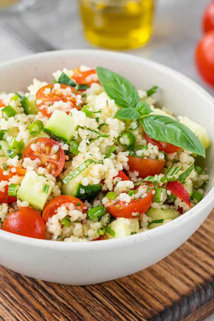 Couscous salad with fresh vegetables and herbs in a bowl on a gray concrete background.