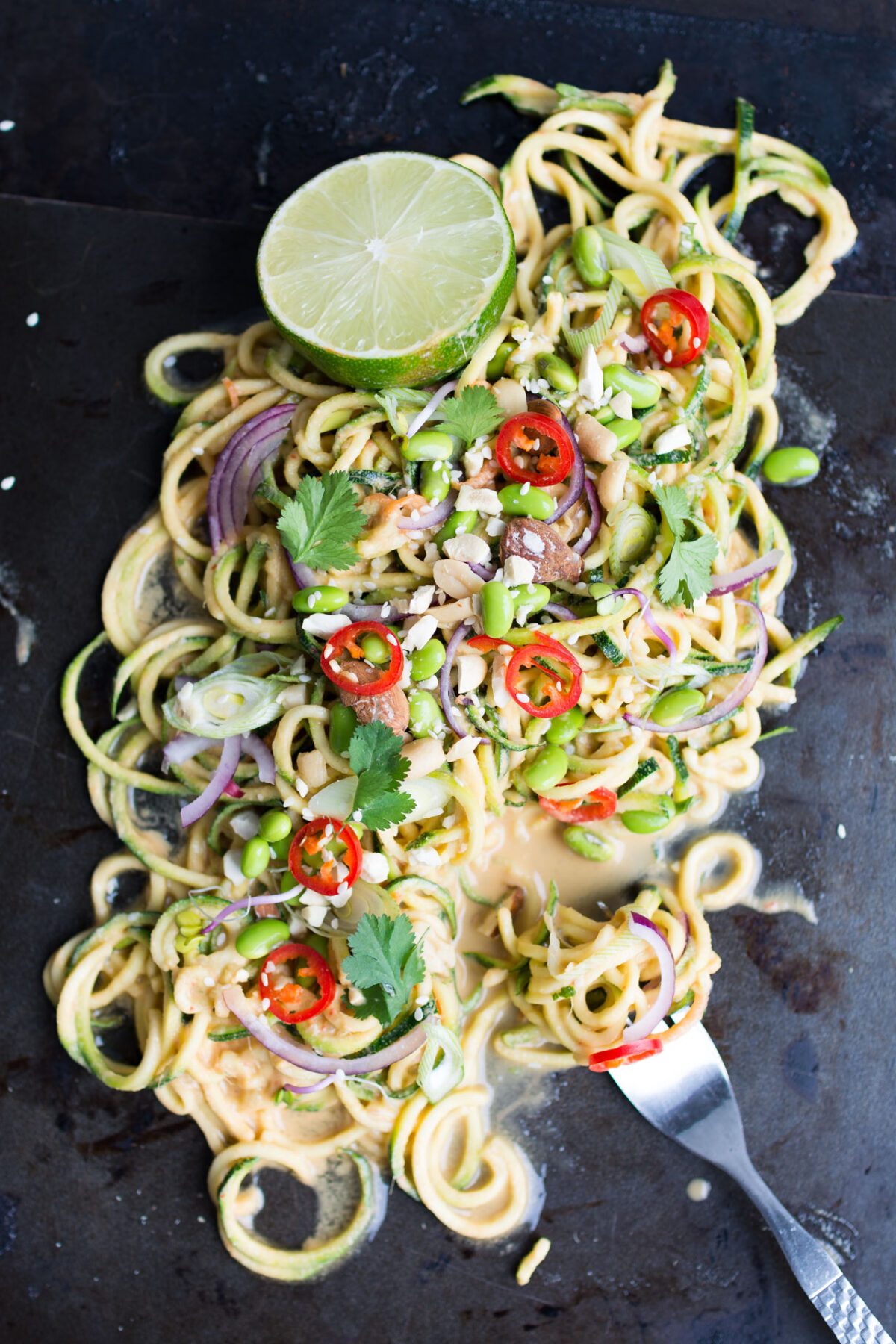 top view image of a thai zucchini noodle salad with fork, top with edamame beans, onions, chopped red chili, peanuts, spring onions, parsley, and sesame seeds