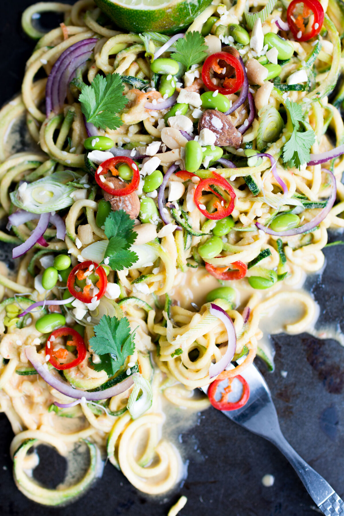 close up view image of a thai zucchini noodle salad top with edamame beans, onions, chopped red chili, peanuts, spring onions, parsley, and sesame seeds